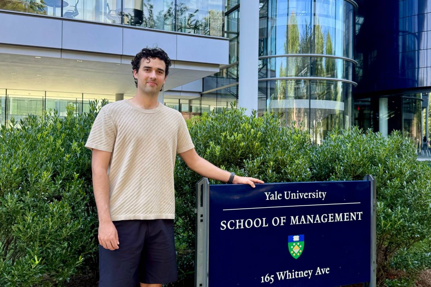 Shane Haynes standing with a hand on the Yale School of Management sign, in front of the school's glass building