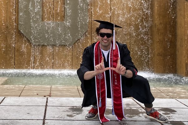 Shane Haynes in cap and gown crouching in front of the block U fountain, making the U sign